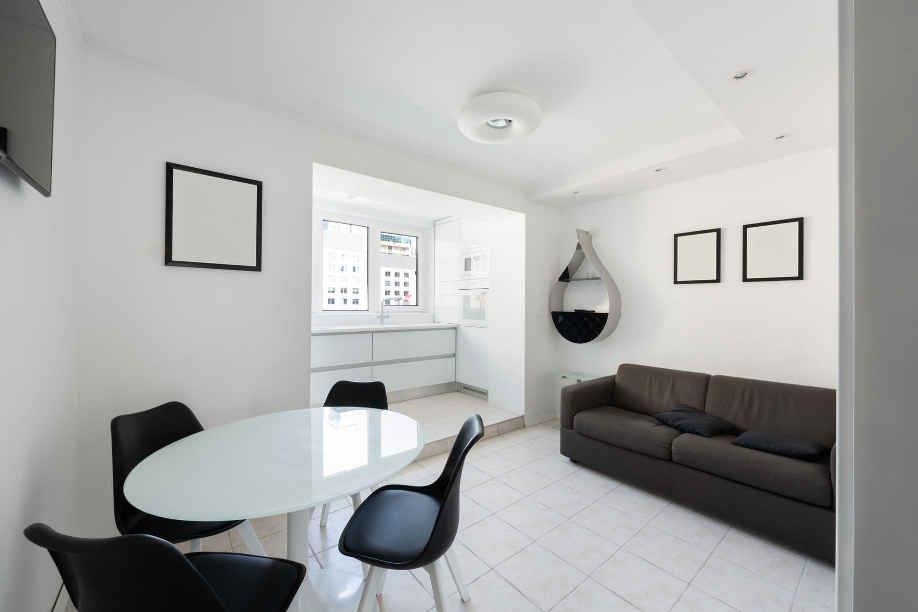 Interior of modern light house with glass table near chairs and couch with pillows near paintings and shelves on wall near entrance to kitchen with counter with window