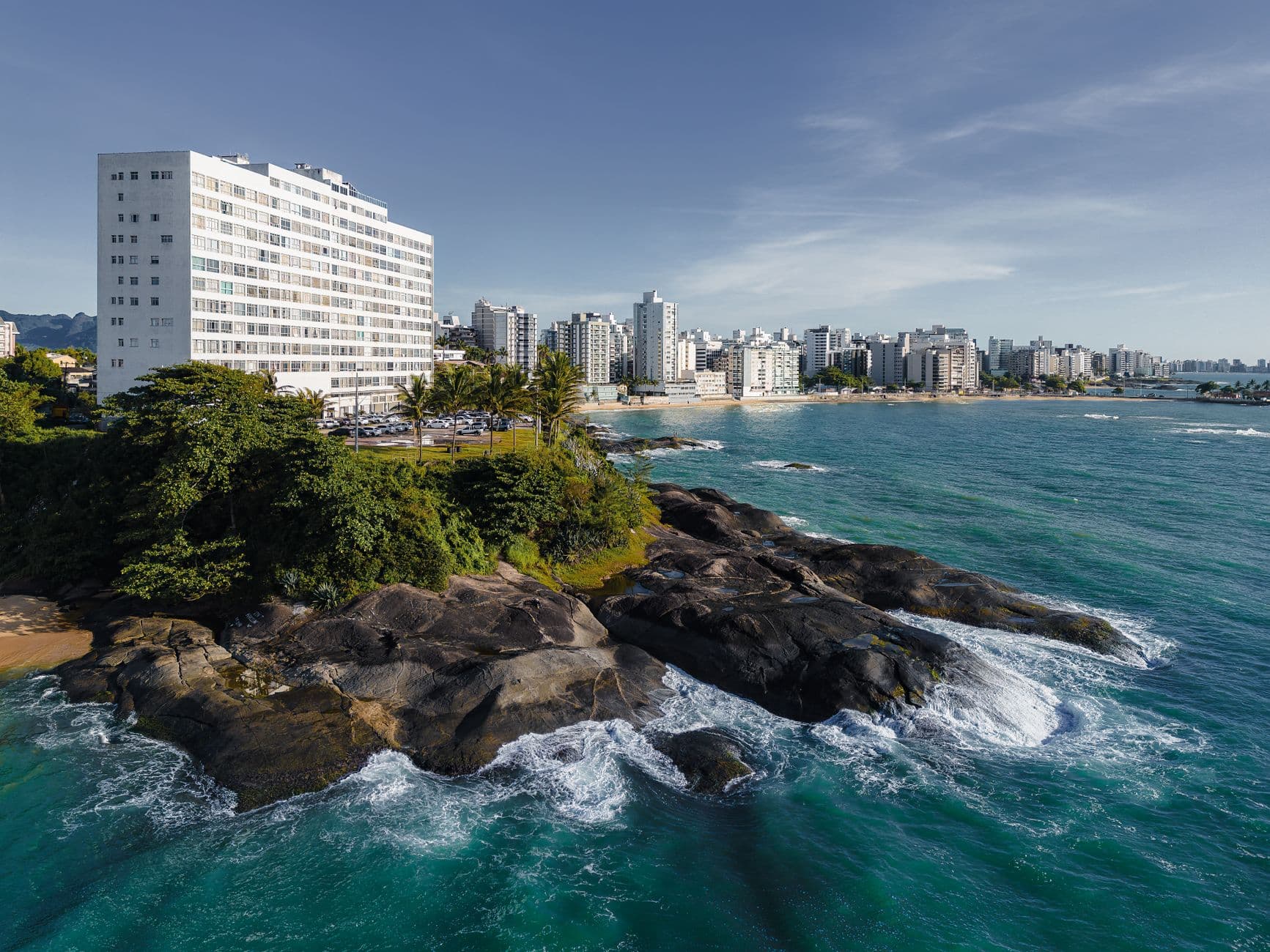 Aerial view of Guarapari's stunning coastline with hotels and rocky seashore under a clear sky.