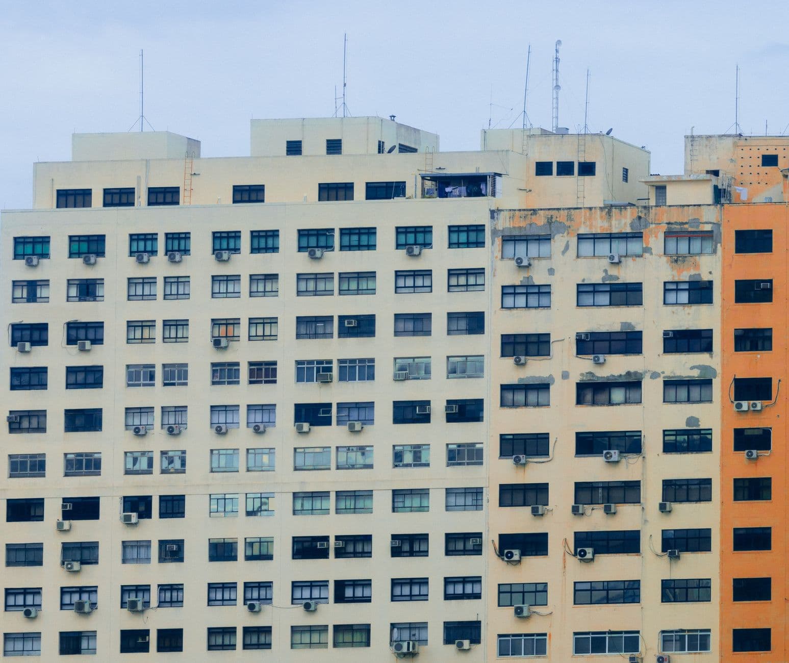 A modern high-rise apartment building with numerous air conditioners and antennas on a cloudy day.